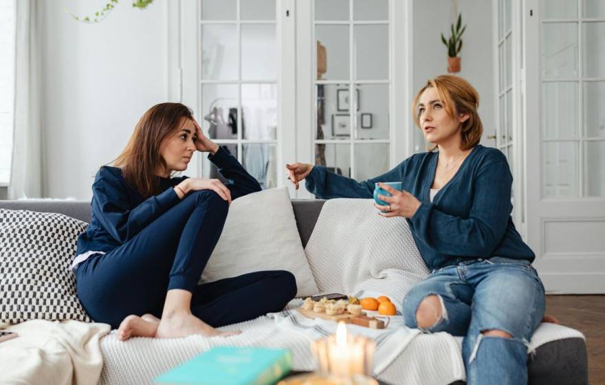 Two women sit on a couch drinking tea with a snack tray between them. The woman on the right is speaking and the woman on the left is listening.