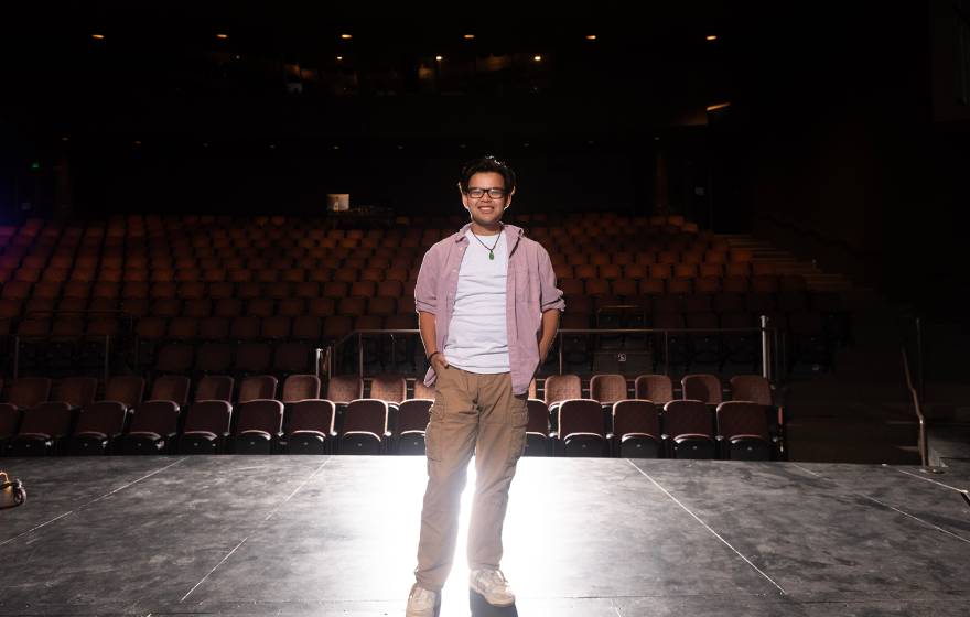 A smiling man standing on an empty theater stage under dramatic lighting