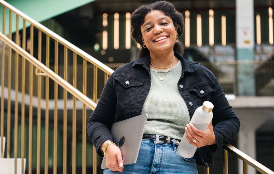 A college student smiles for the camera on a staircase in a university building, holding a reusable water bottle and a laptop