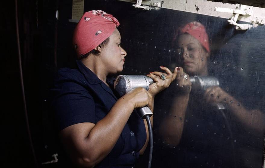 A Black woman uses a rivet gun on a reflective steel panel, wearing a red headscarf and a blue jumpsuit,