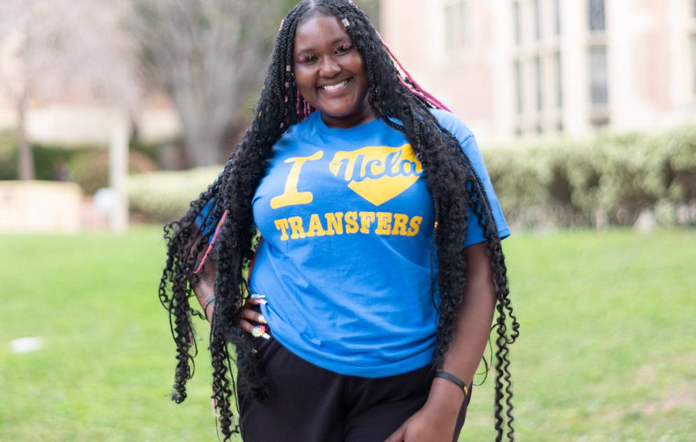 A woman with long braids and an I heart (UCLA) transfers shirt smiles on the UCLA campus