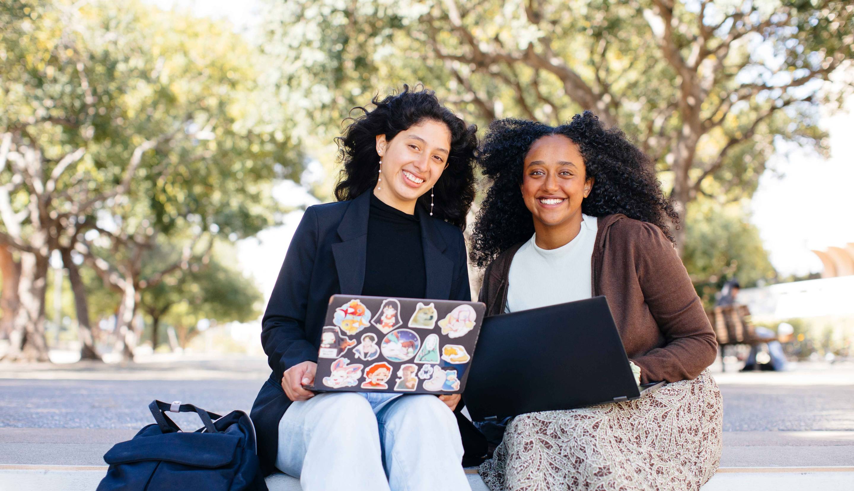 Two women smiling while holding a laptop on campus. 