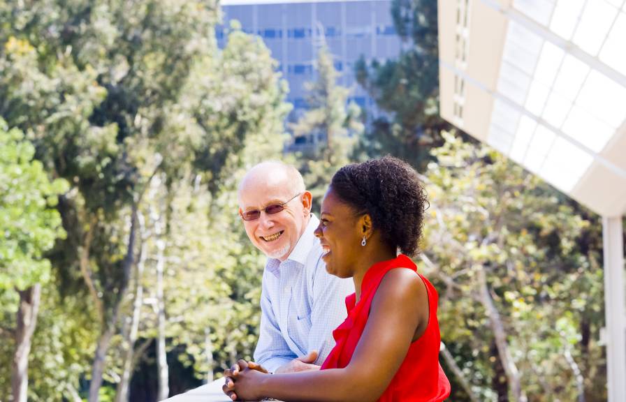 An older white man and a young Black woman in a dress talk outside on a leafy campus
