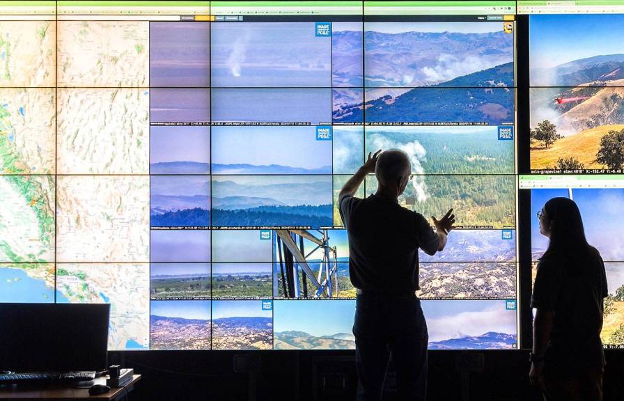 Two people in silhouette in front of a giant screen with many colorful CCTV video monitors overlaid on a map of california