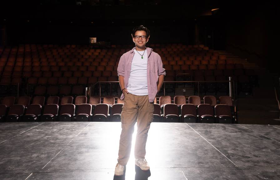 A smiling man standing on an empty stage under dramatic lighting