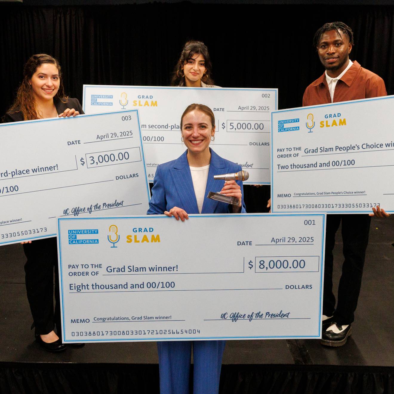 Grad Slam 2025 winners Four people, one in the foreground, three in the background, hold up giant checks