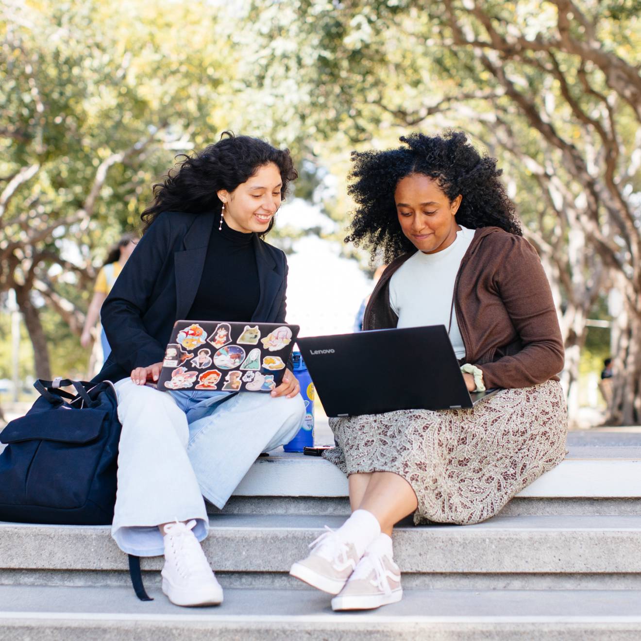 Two students sitting on a stairway outside looking at laptops together and smiling