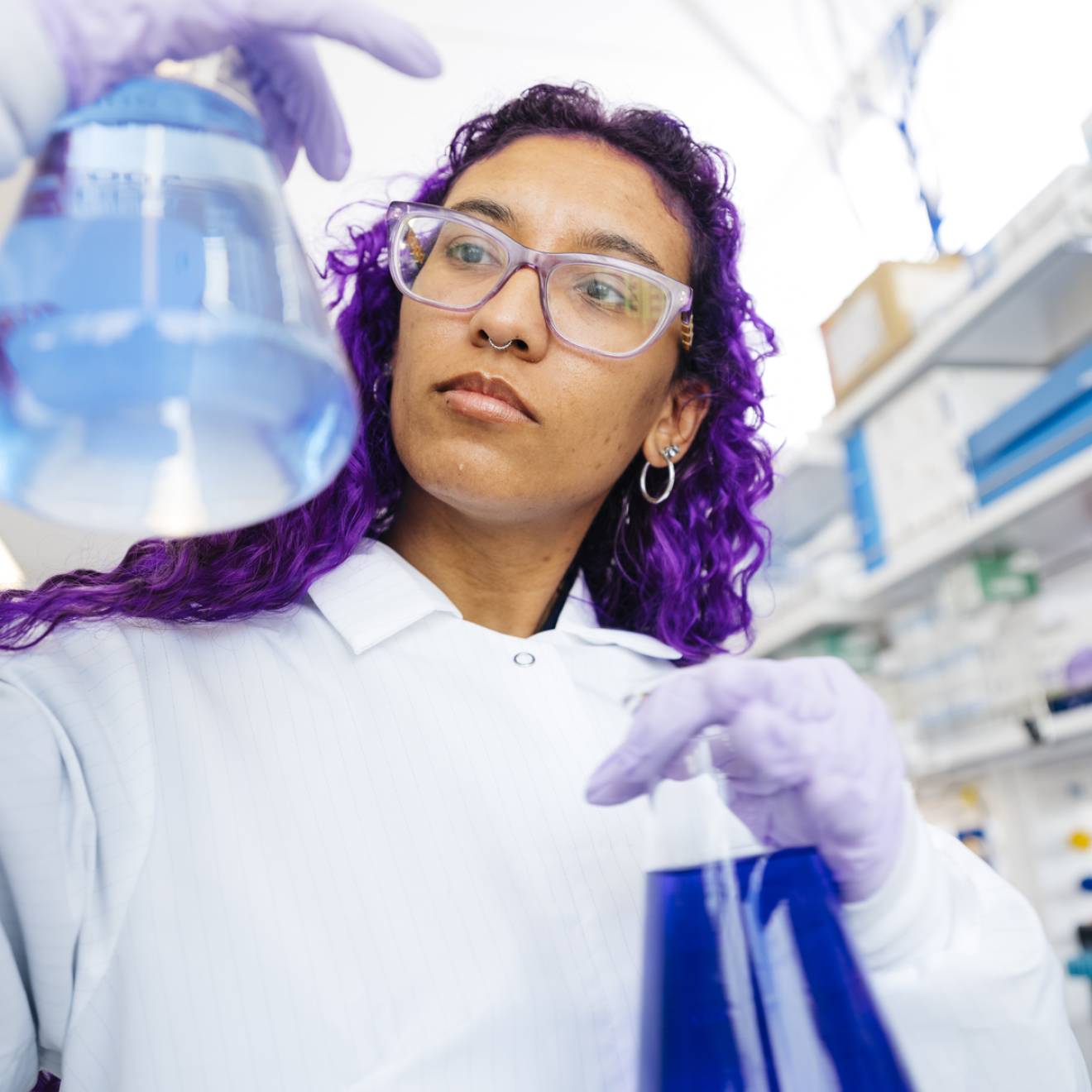 Woman with long, dyed-purple hair wearing a white lab coat and protective gear compares two flasks full of colorful liquid in a lab