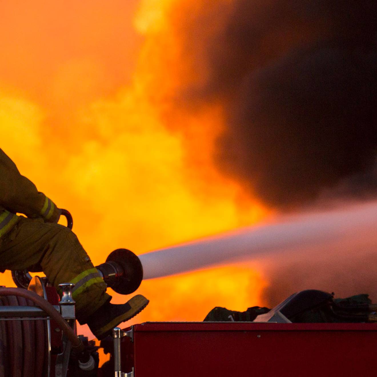 A firefighter straddles a roll of hose, aiming a jet of water into a wall of flames, facing away from the camera