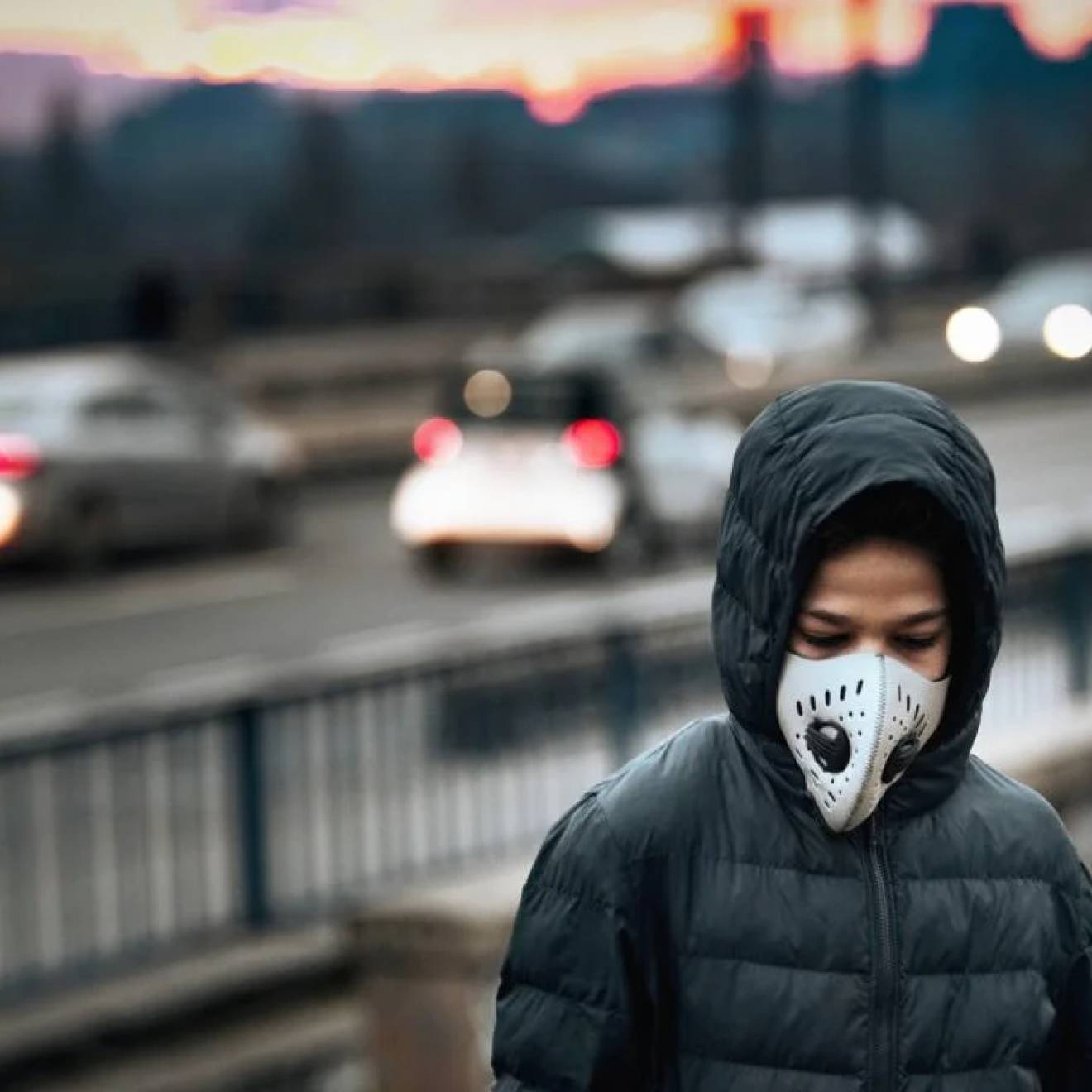 A boy in a hooded jacket standing next to a freeway wearing a respirator mask