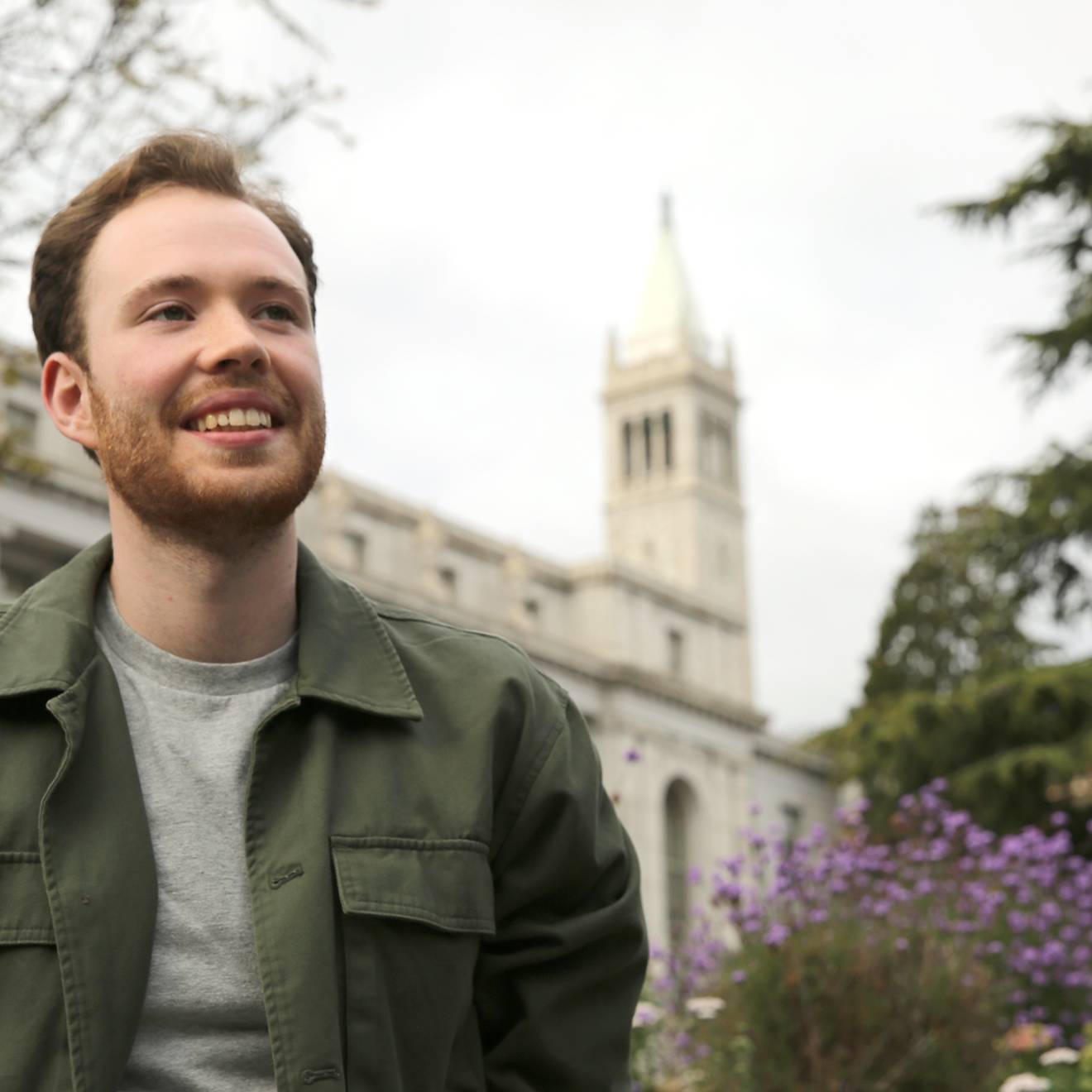 Asher Cohen, University Medalist a young man wearing a green jacket puts his hands in his pockets and smiles in front of the Campanile