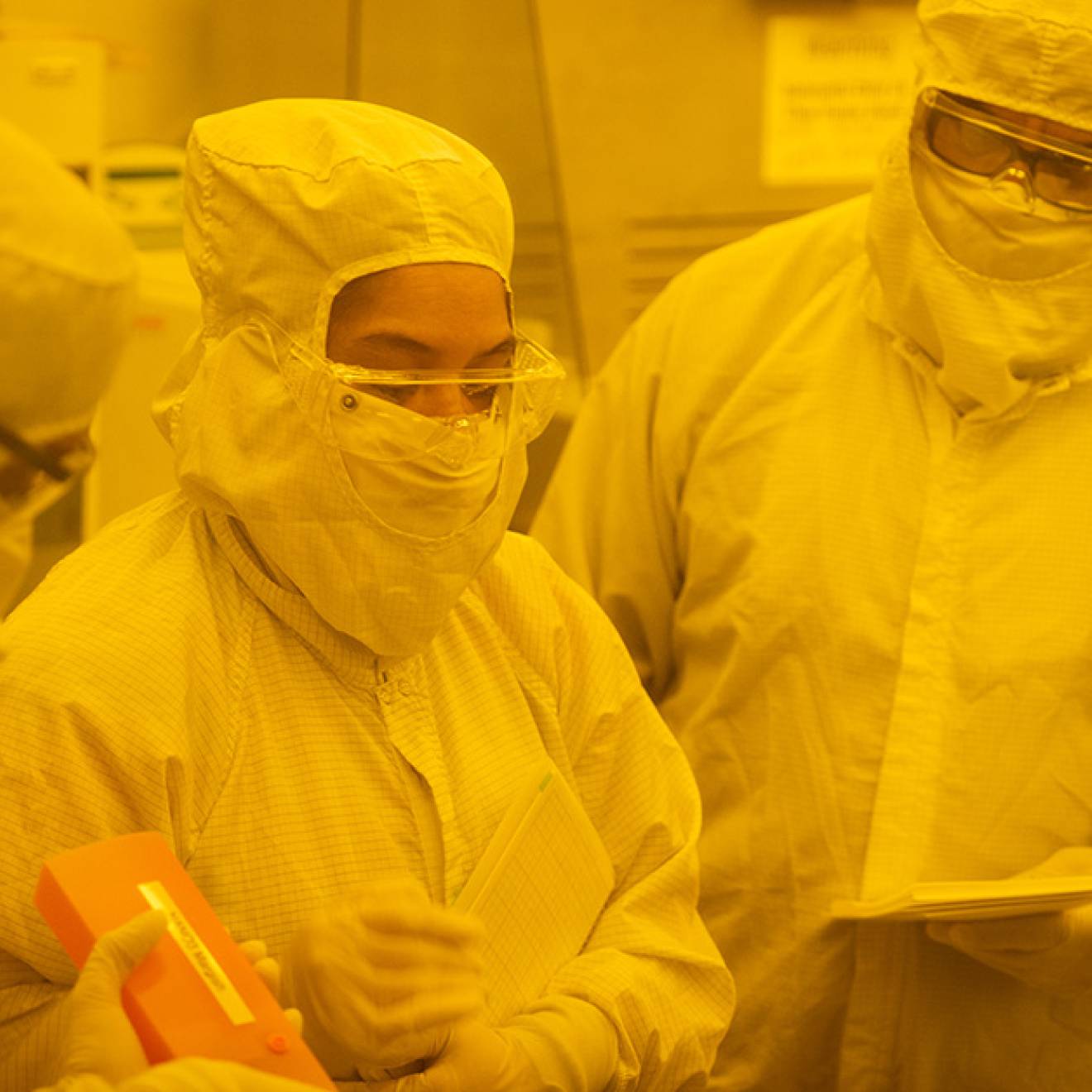 Three people stand in white full-body suits in the yellow light of Nano3's cleanroom.