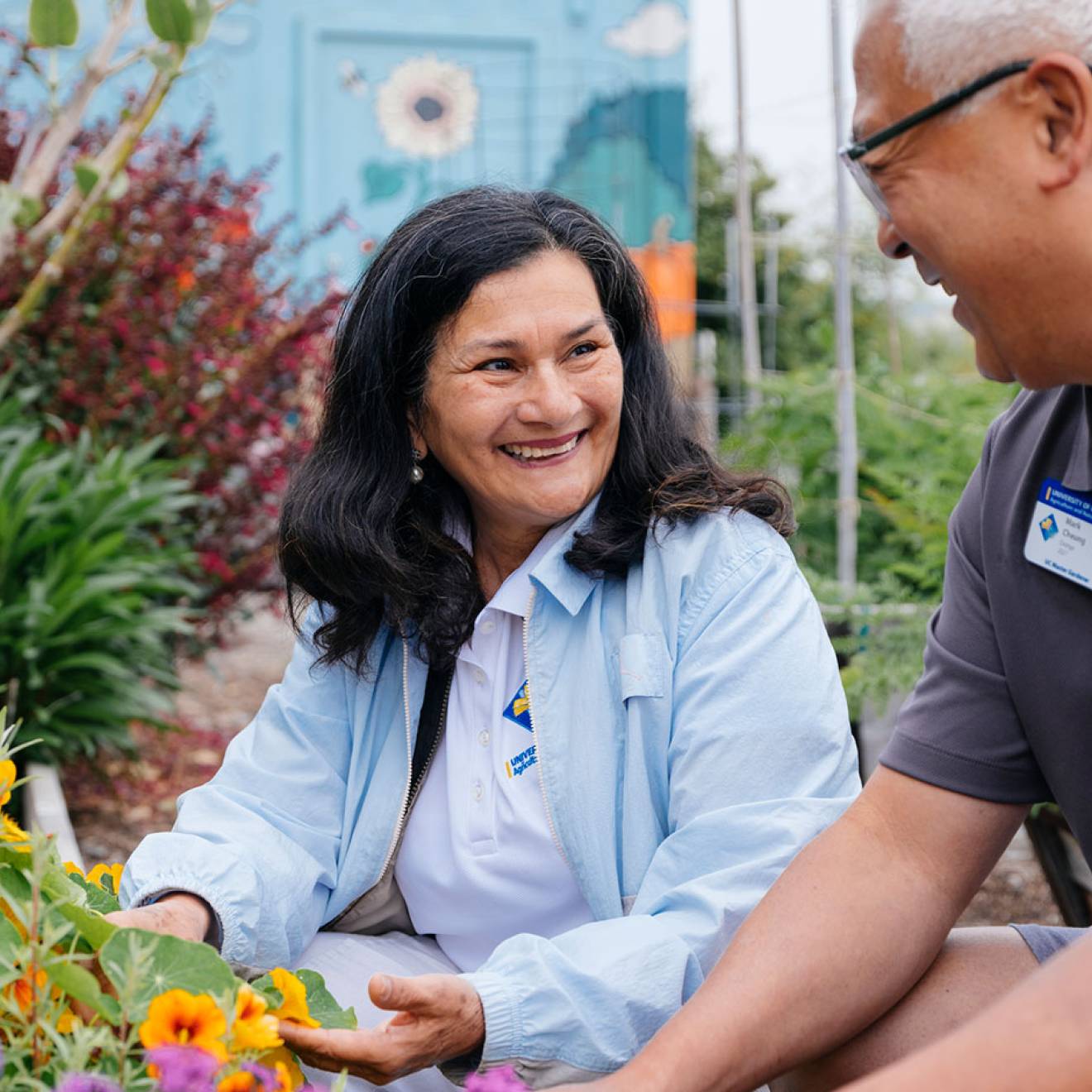 A man and woman smile while tending to a colorful garden.