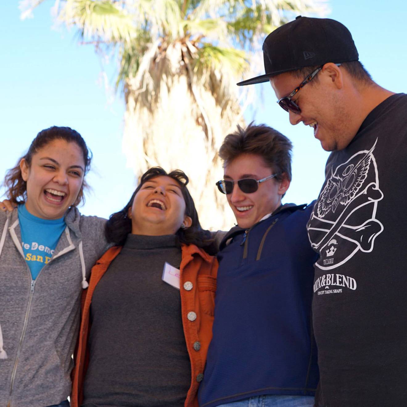 Four students with arms around each other laugh and smile, with a palm tree in the background