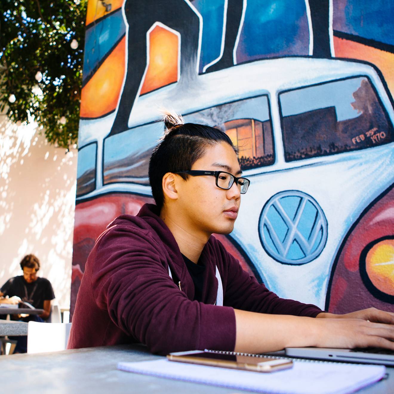 A student working on a laptop at a table outside, in front of a mural showing a Volkswagen van