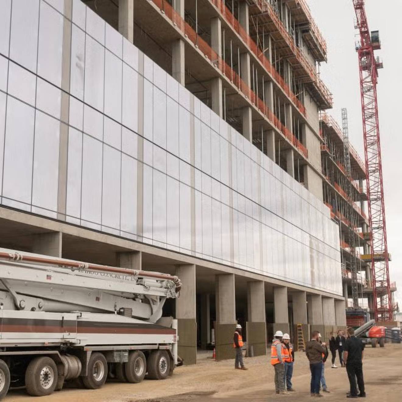 A building  with numerous glass panels under construction on UC Davis' Aggie Square with a construction crew talking below it next to a truck with a crane