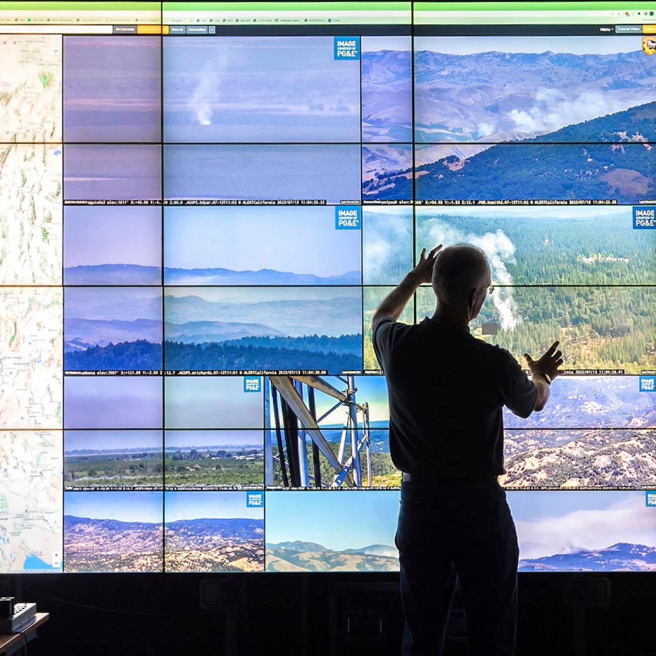 Two people stand in front of a wall of screens showing a grid of footage of natural landscapes, a map of California and some smoke from a wildfire 