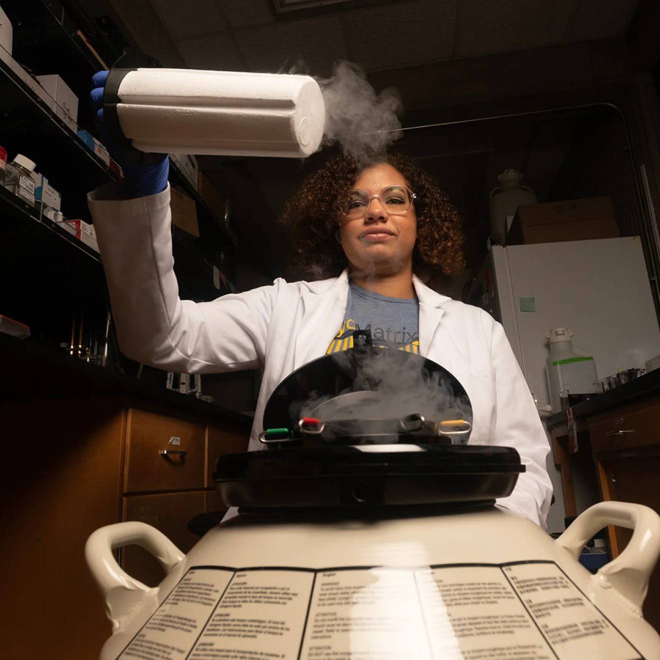 A young woman graduate student with curly hair pulls out a nitrogen storage cylinder and holds it over a piece of equipment