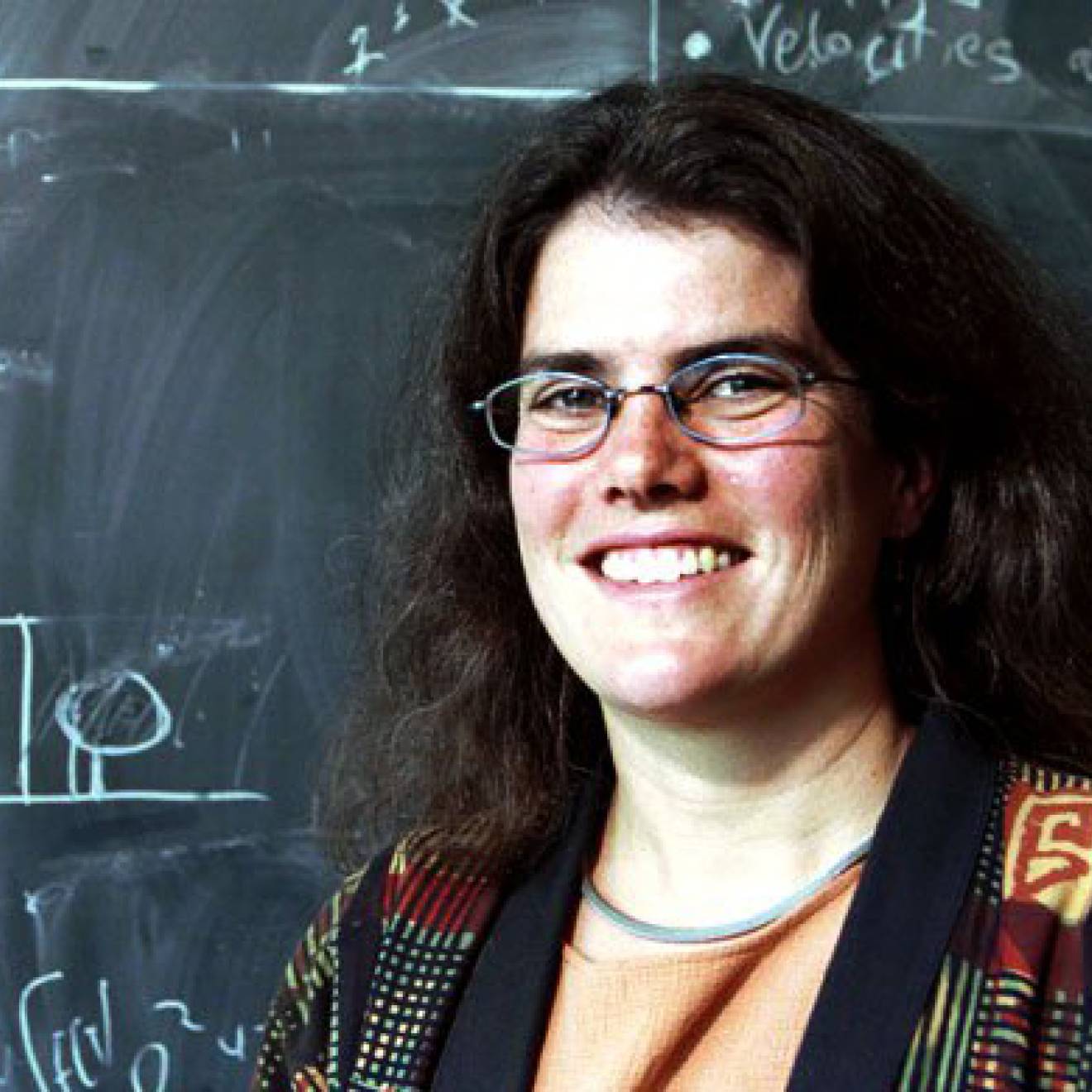 A woman wearing glasses standing in front of a chalkboard covered in scientific notations