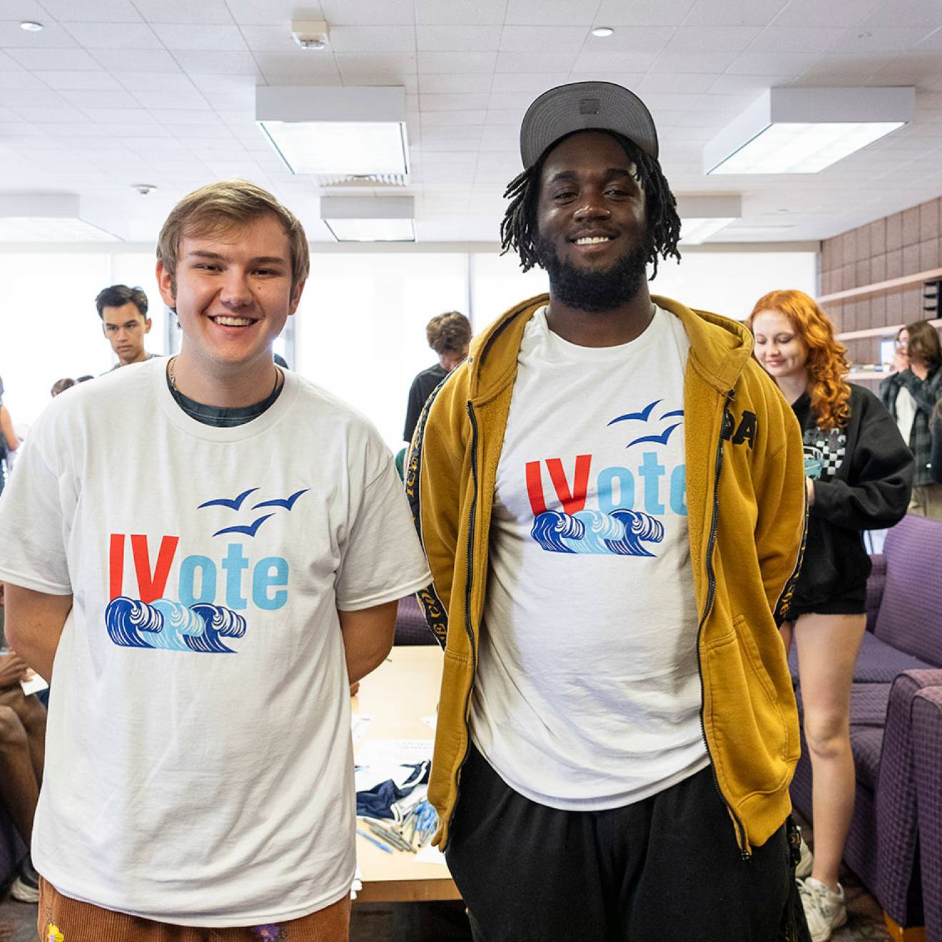 Two UC Santa Barbara students face the camera and smile, wearing t-shirts reading "IV Votes", standing in a busy student lounge.