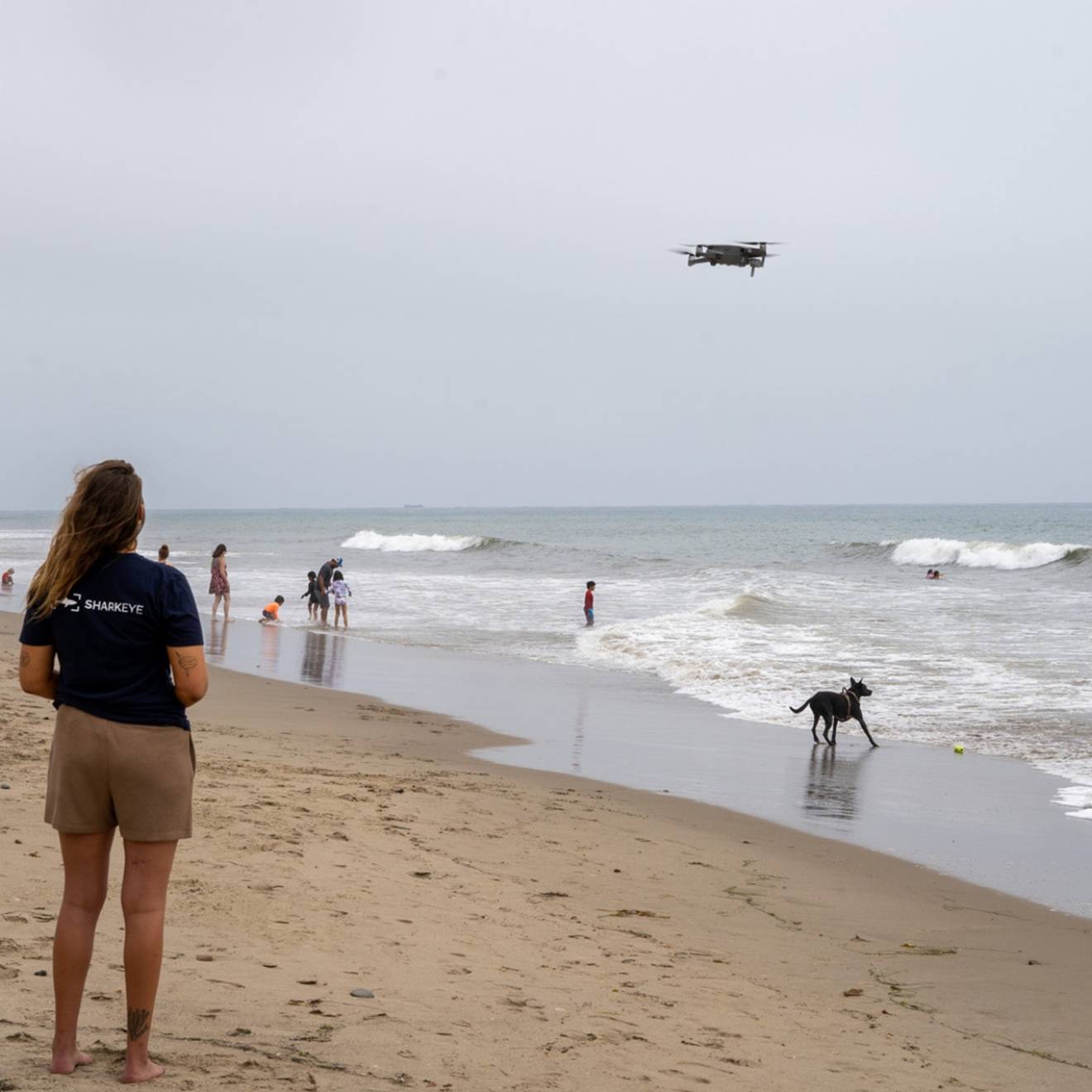 a woman stands on a busy beach looking out a a drone hovering above the waves.