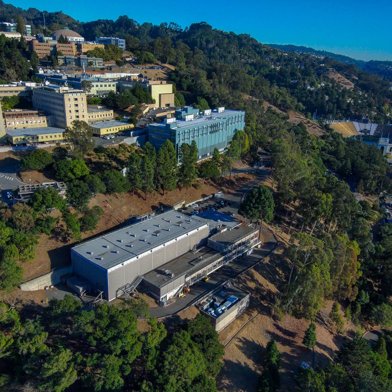 A number of buildings on a tree-covered hillside, including Berkeley Lab, seen from an aerial view on a clear day, Bay in the background