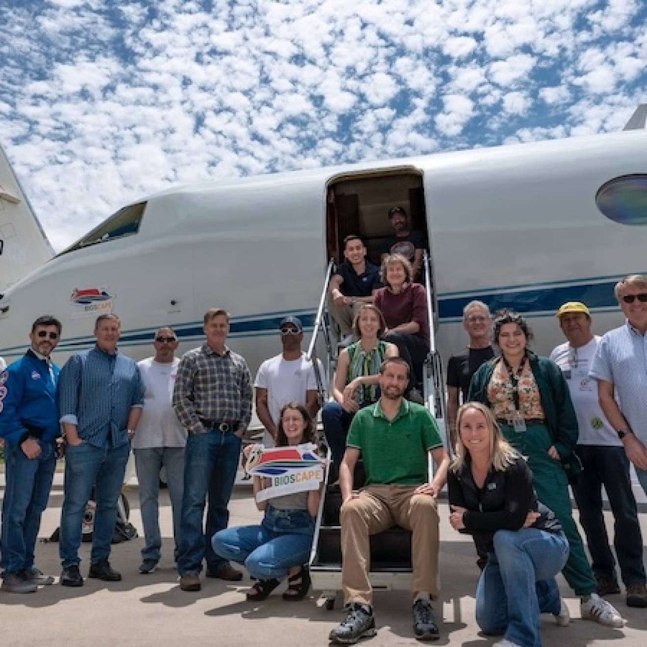 About 20 people gather for a photo on an airport tarmac, in front of a small jet