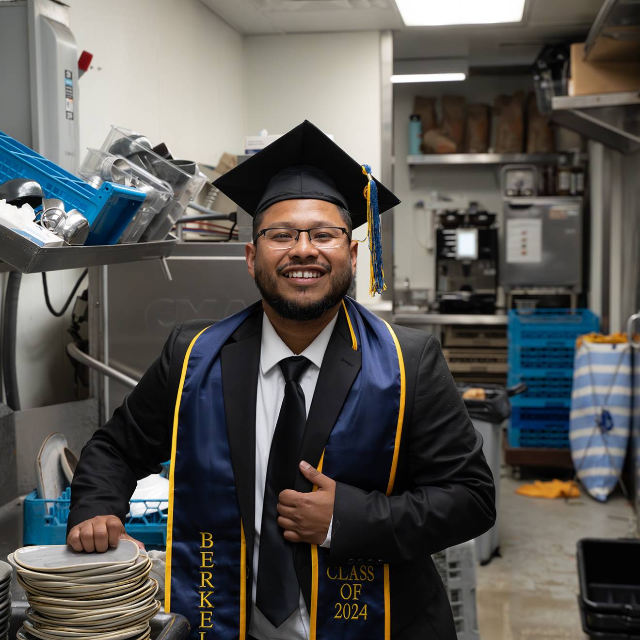 A smiling man in a college graduation cap and gown with a UC Berkeley sash stands in a commercial kitchen in front of a sink and a stack of dishes