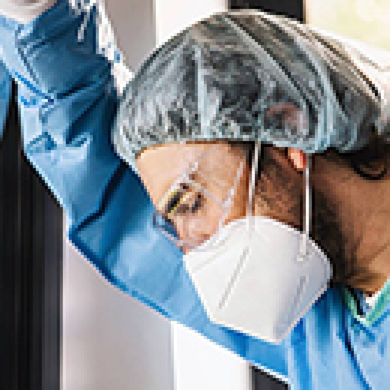 A man in scrubs, a hairnet and a face mask leans against a window