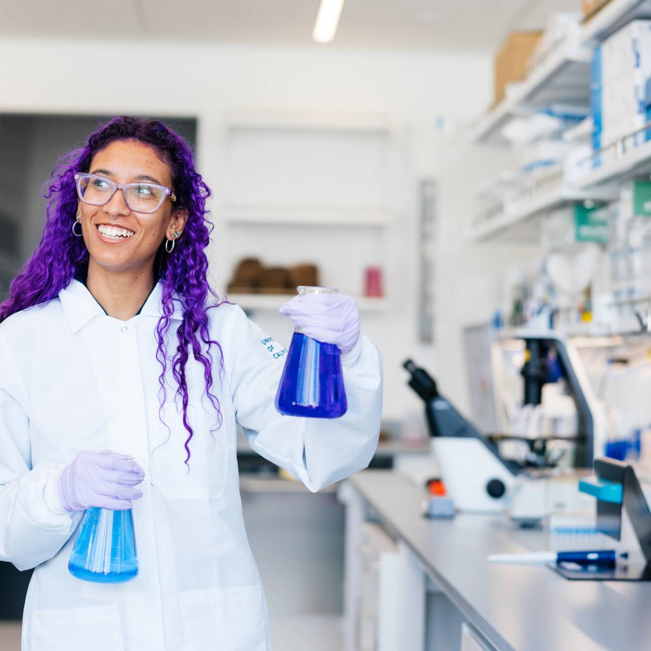 A woman with two flasks of blue chemicals and long purple hair in PPE and a lab coat smiles at a lab table