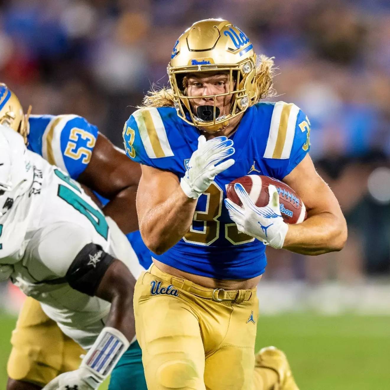 A player in UCLA blue and gold carries the ball and runs by players in white uniforms