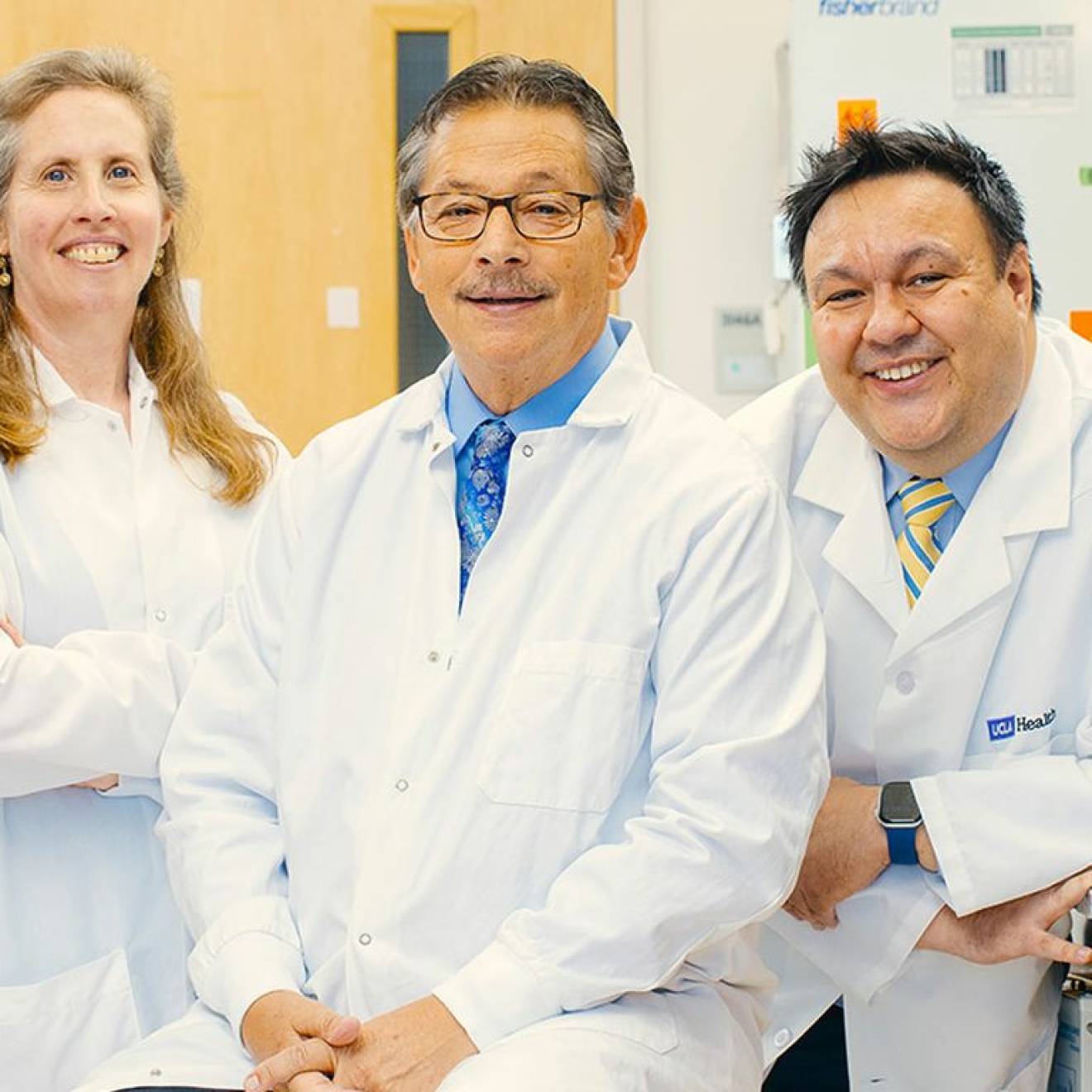 Three doctors with white lab coats pose together in a lab and smile