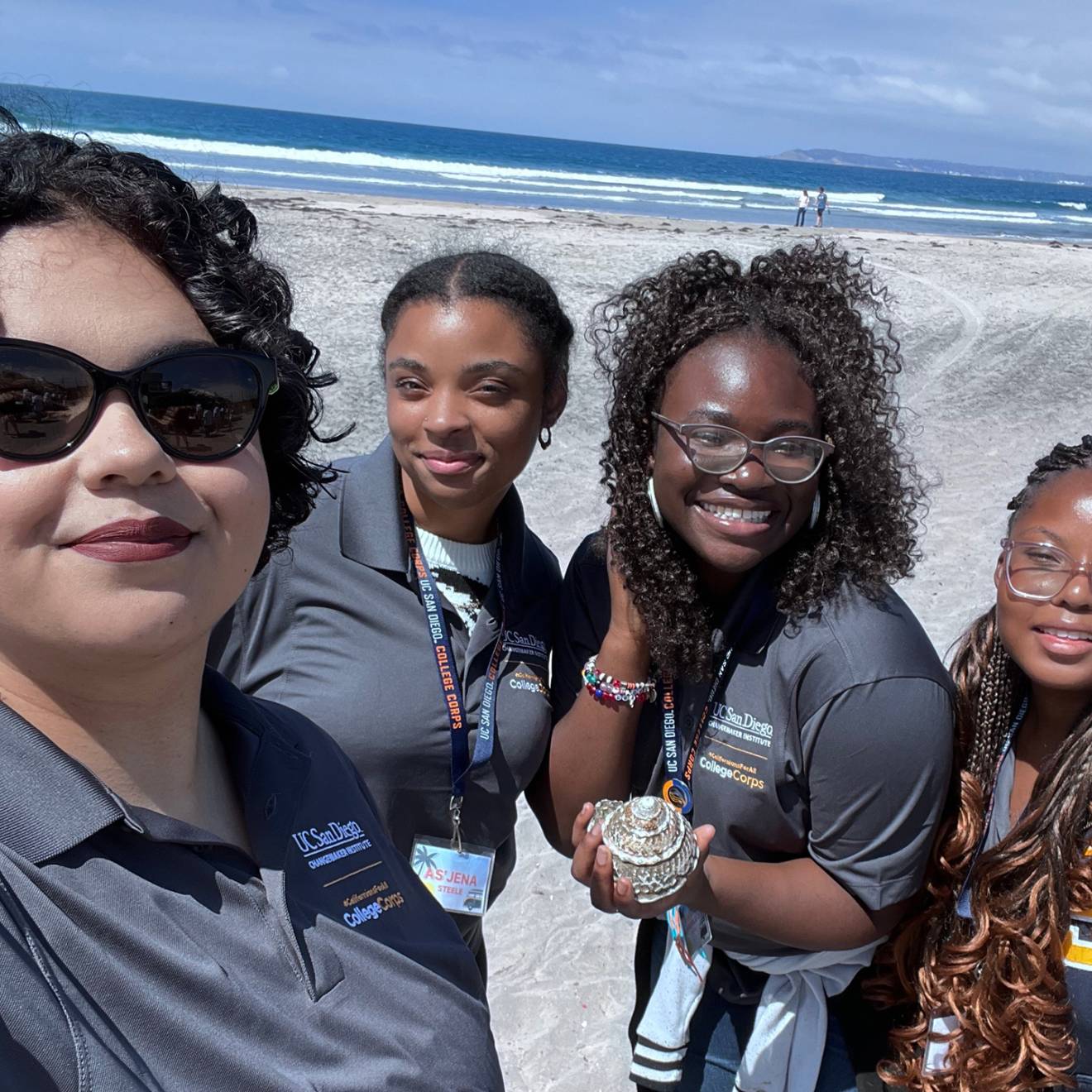 College Corps Four women in gray UC San Diego College Corps polo shirts take a selfie on the beach, smiling