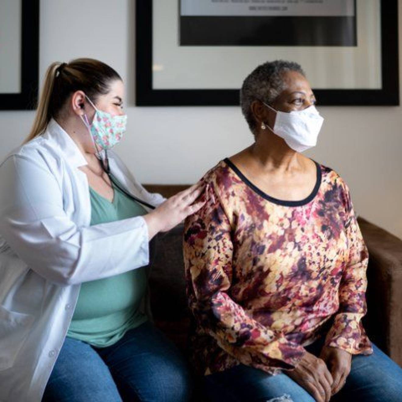 Doctor examining an older Black woman's breathing, both wearing masks