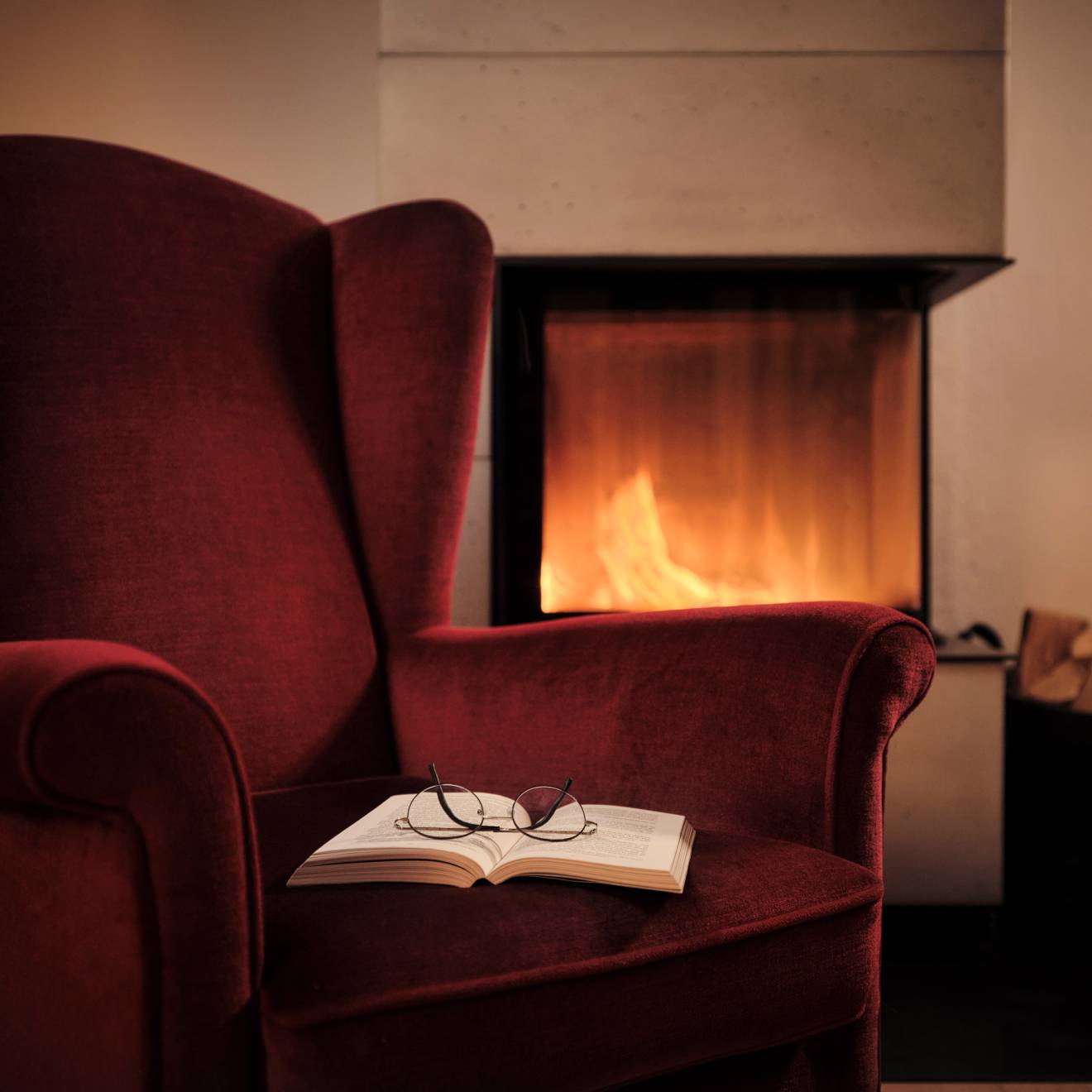 A deep red arm chair with an open book with glasses on top of it next to a fireplace