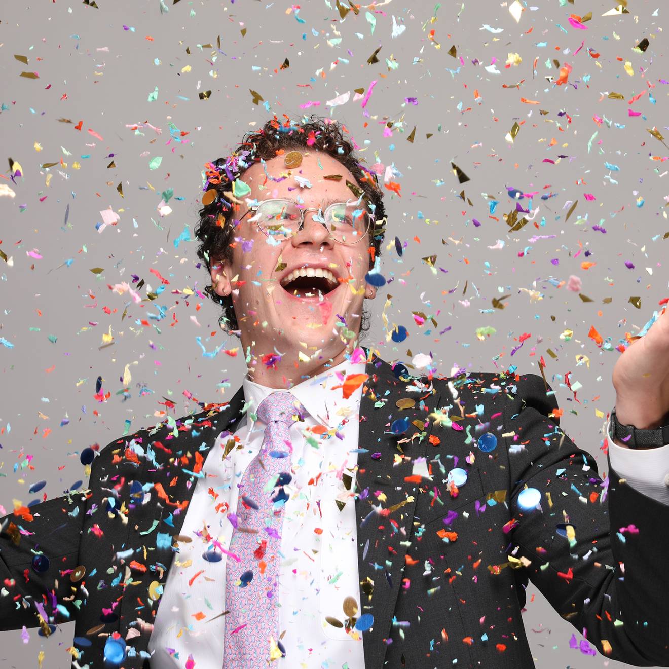 Spelling bee list-maker An ecstatically smiling man with glasses in a suit with a pink tie holds up his arms while confetti falls