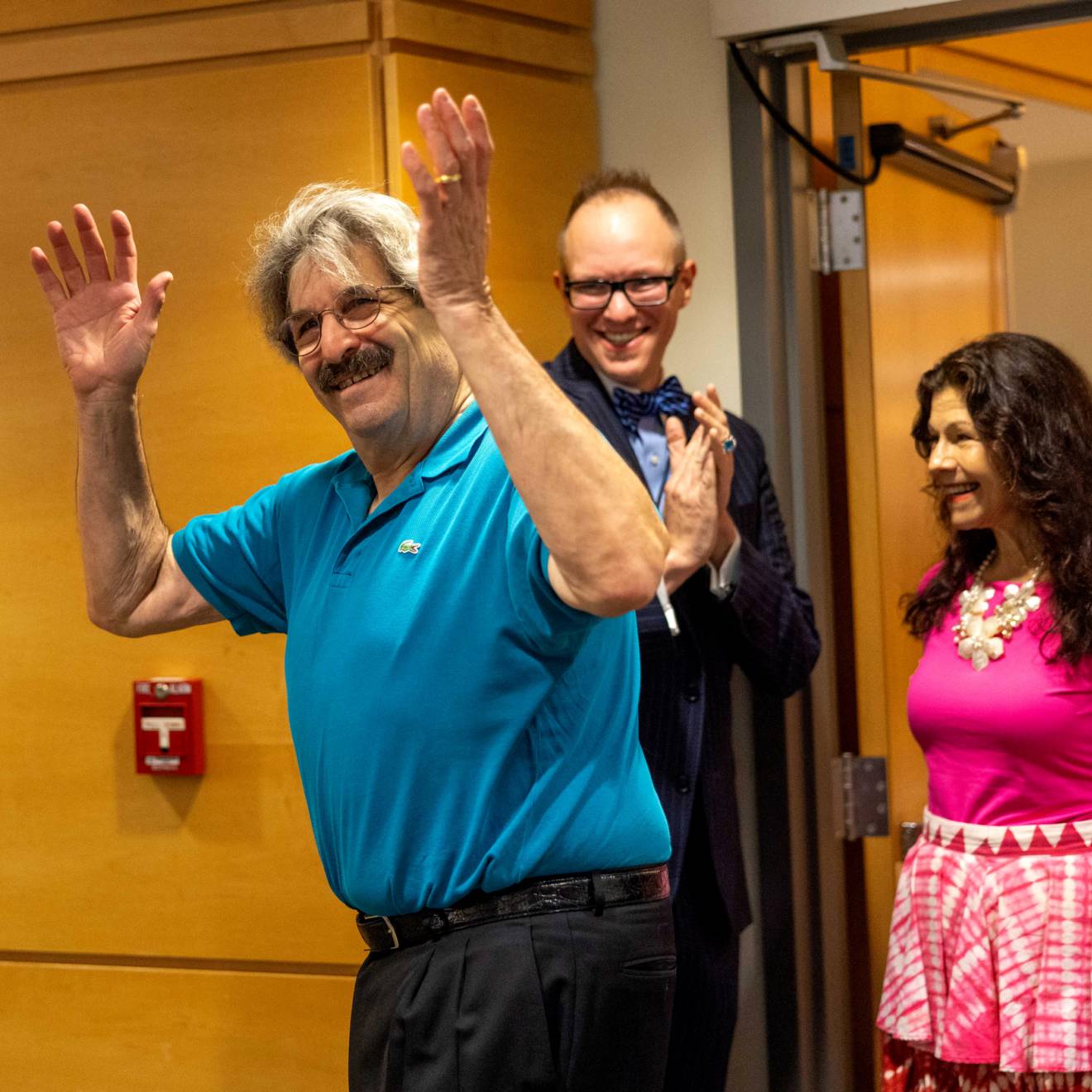 An older man with thick hair and a mustache and glasses throws up his hands as he enters a room and smiles as a woman in a pink dress follows behind, also smilling, and a man behind them smiles, looking on