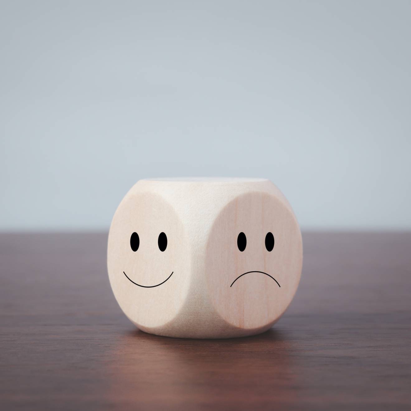 A 6-sided die on a wooden table top. Of the 2 faces visible, one has a happy face and the other has a sad face drawn on it