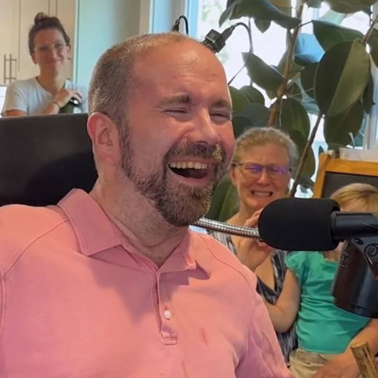 A man with a beard and a pink shirt in his living room grinning, overjoyed in front of a microphone, while his family watches