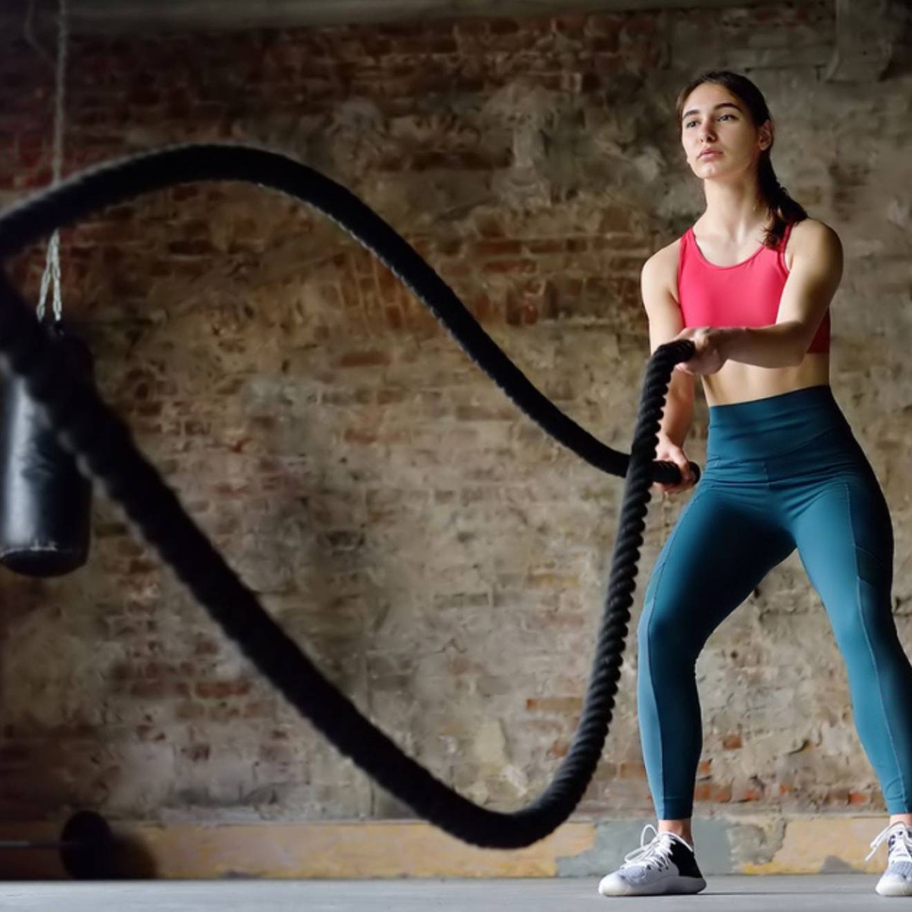 Woman in athletic attire uses battle ropes in a room with an exposed brick wall