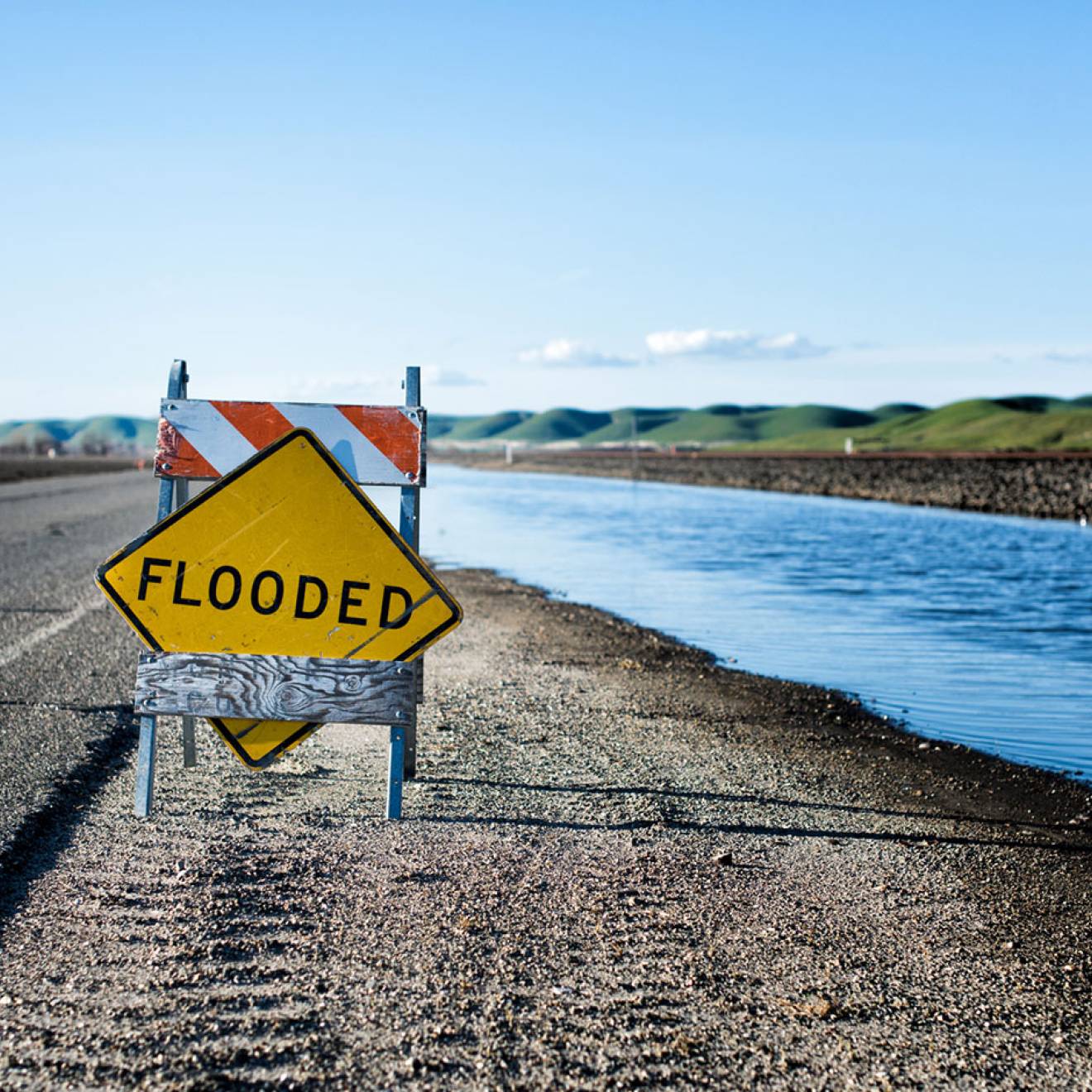 A temporary road sign reads "FLOODED", placed on a highway shoulder next to a canal, with green hills in the background