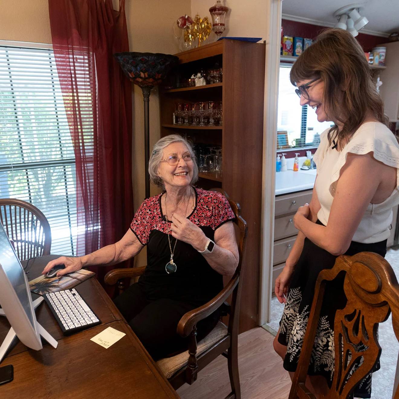 An older woman with gray hair and glasses sits at a computer at the dining room table, smiling and talking with a younger woman standing beside her