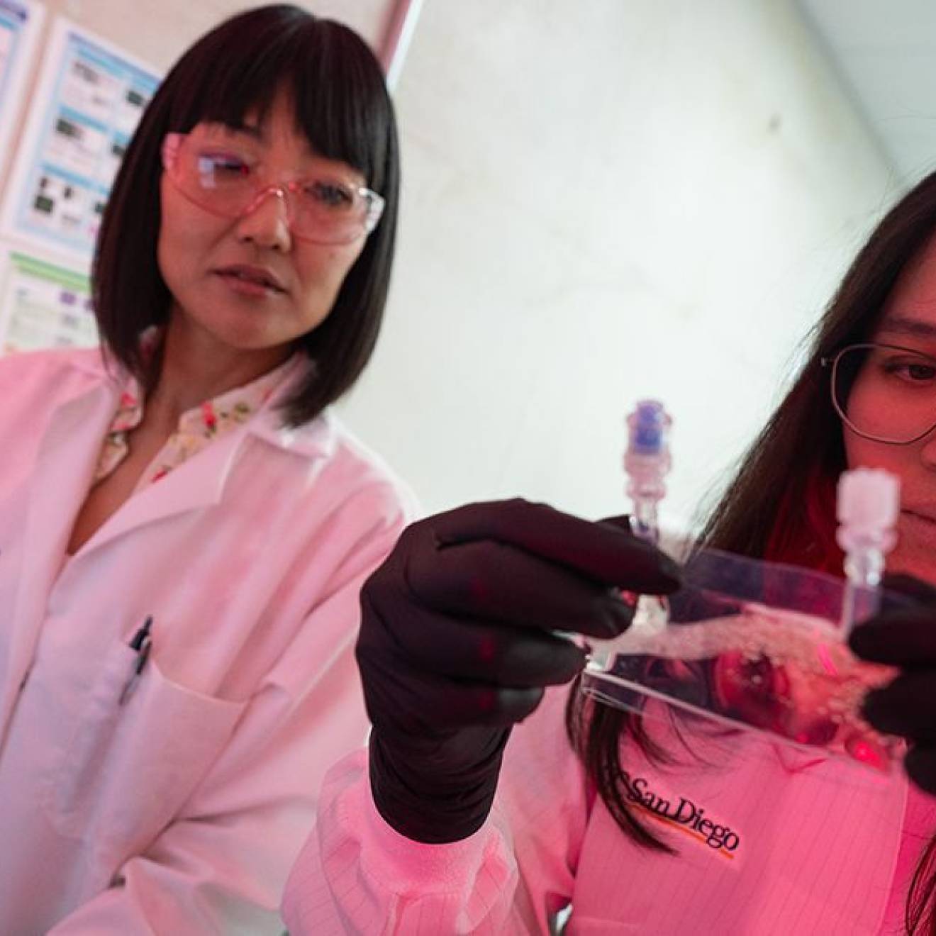 Cancer stem cells Two women in lab coats in a lab with reddish lighting in the corner; one holds and looks at a small baggie