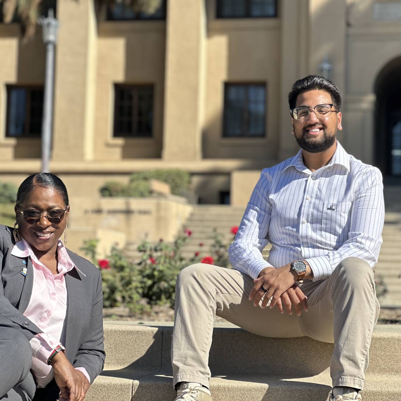 Young Black woman in a gray suit with pink blouse and sunglasses, smiling (Sonya Brooks); and young man (Josiah Beharry) with beard, glasses, khakis, dress shirt, smiling. Both seated on steps at the UC Riverside campus on a bright, sunny day.