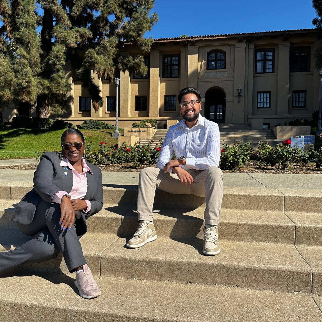 Two student Regents, Josiah Beharry and Sonya Brooks. Sonya is seated on stairs closer to the camera in a business formal gray suit and white blouse, smiling. Josiah is seated higher up on the stairs in a dress shirt and khakis, smilling.