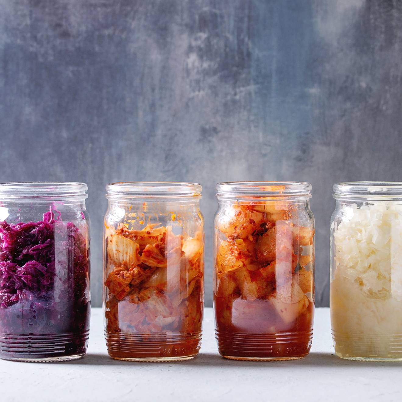 Variety of fermented food Korean traditional kimchi cabbage and radish salad, white and red sauerkraut in glass jars in row over grey blue table