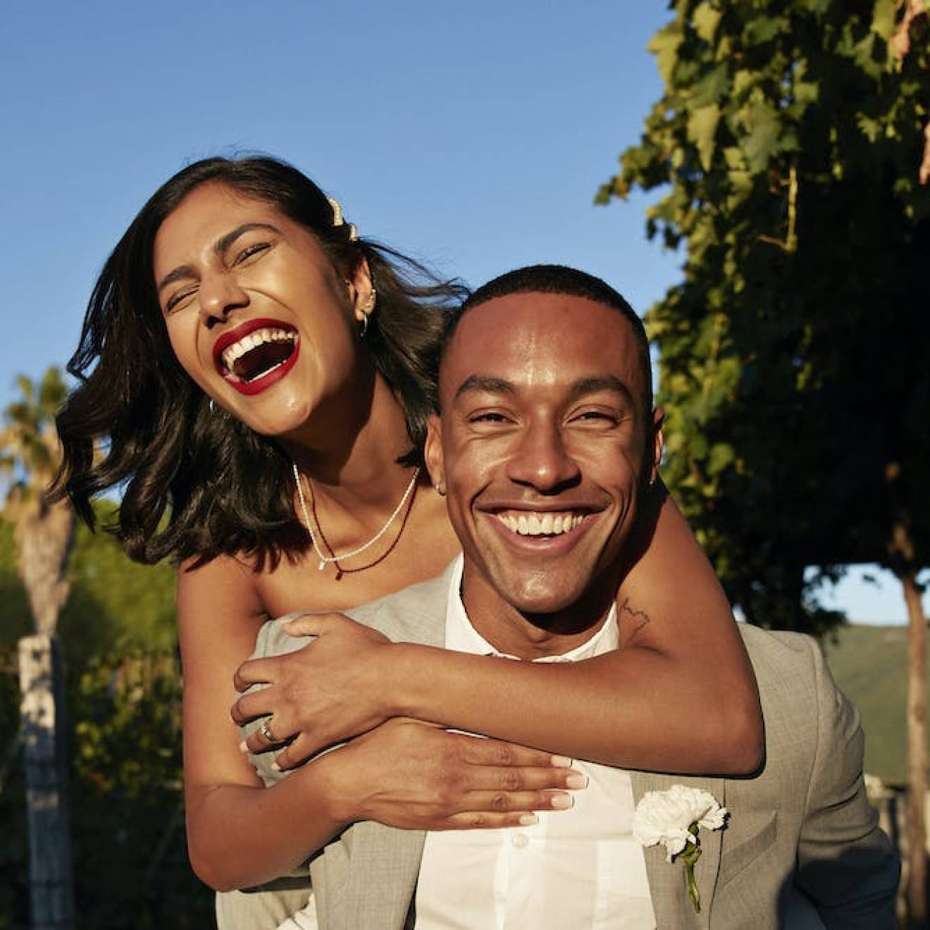 A happy young Latina woman laughing while hanging off the back of a happy young Black man, both in wedding outfits