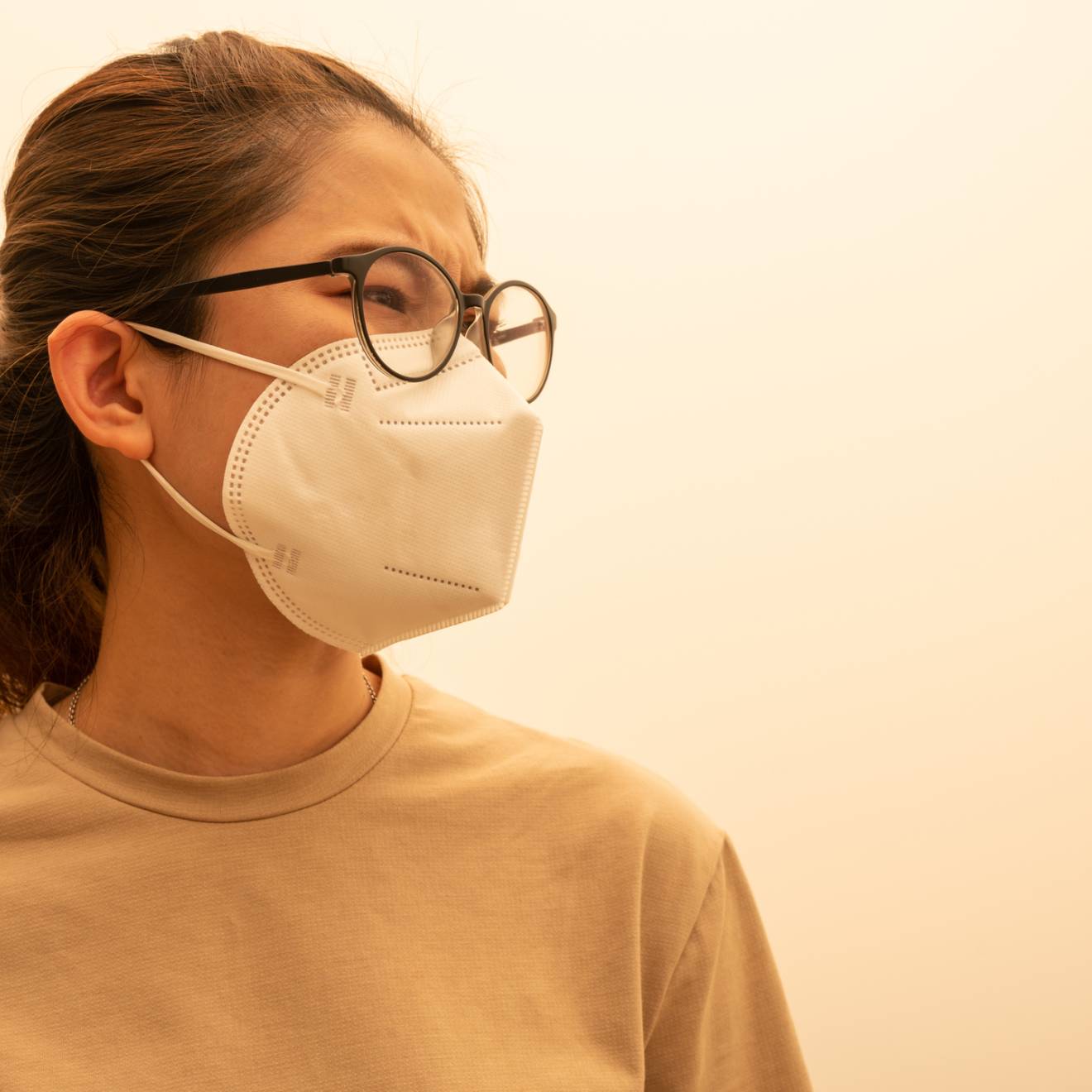 A woman with her hair pulled back wears a N95 mask, looks pensively, yellowish sky around her