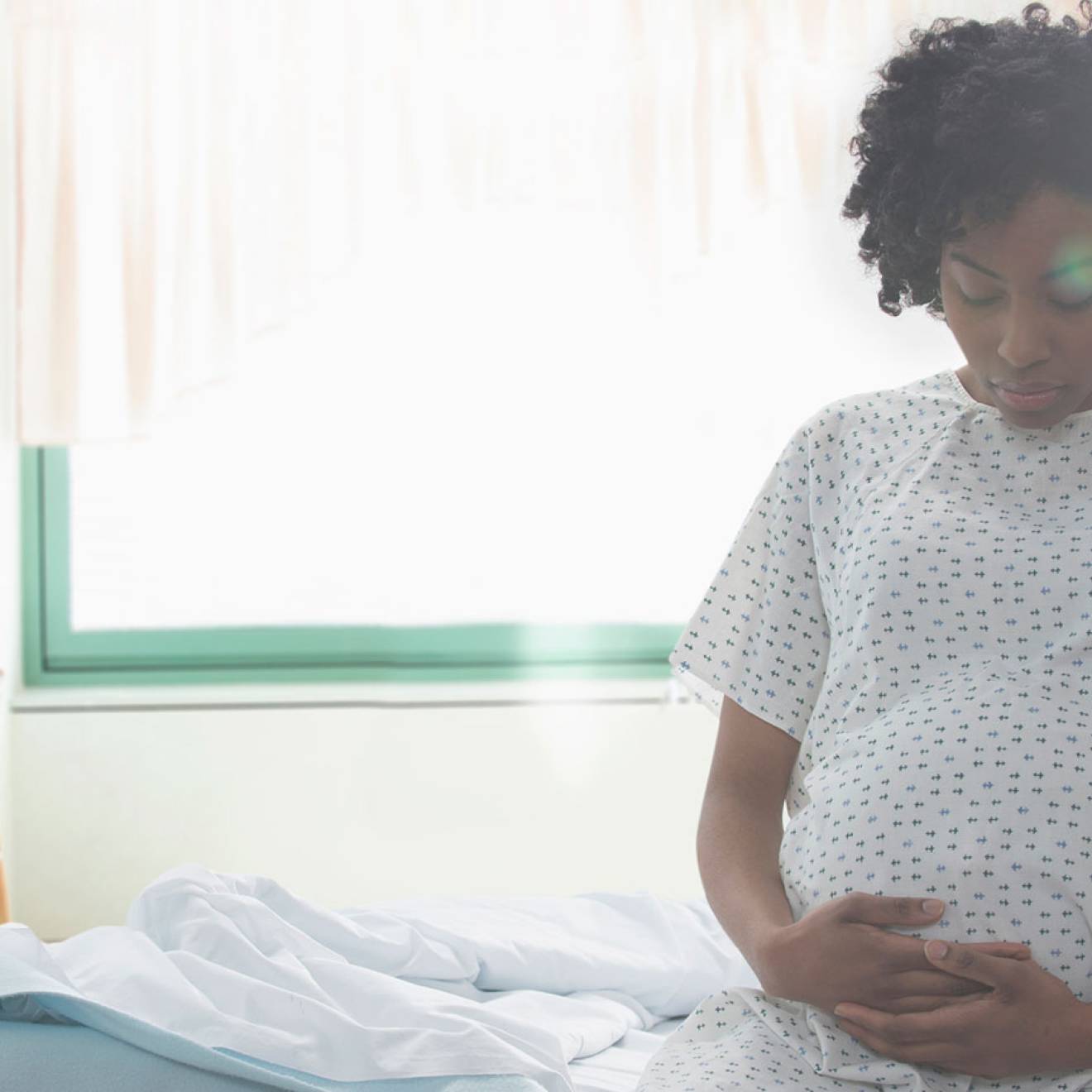 A pregnant woman sits on a hospital bed, wearing a hospital gown, holding her belly from below.