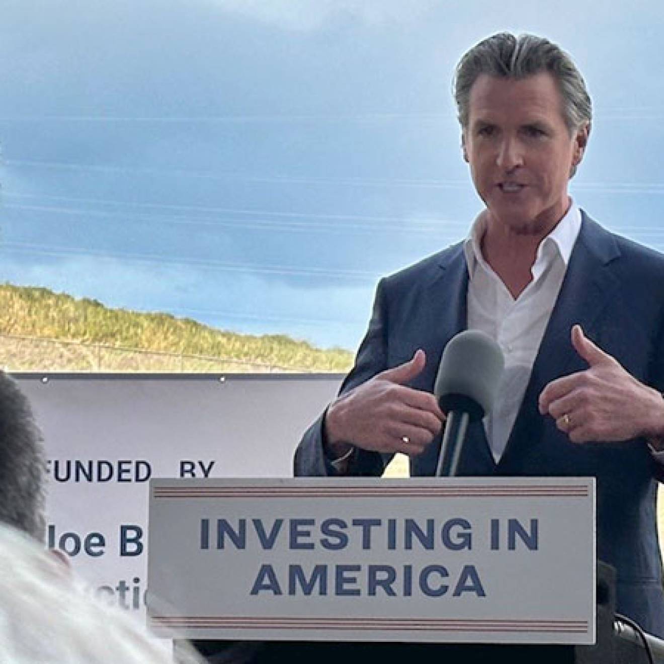 Governor Newsom speaking at a podium that says Investing in America, with countryside behind him